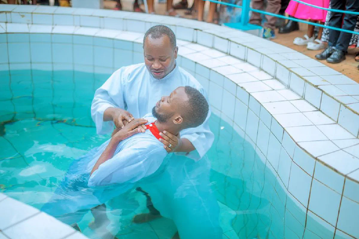A man is being baptized by a priest in a clear water pool, celebrating faith.