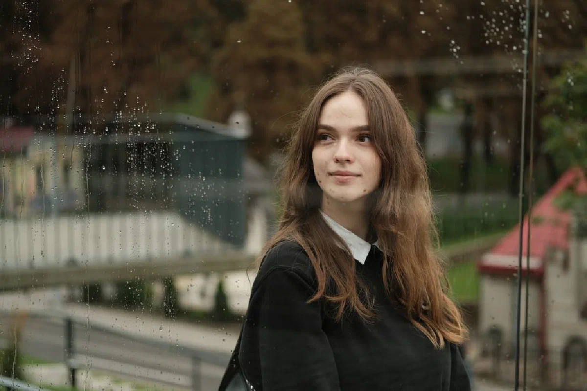Reflection of young smiling female brunette with long dark hair in black sweater standing on street and looking at reflection