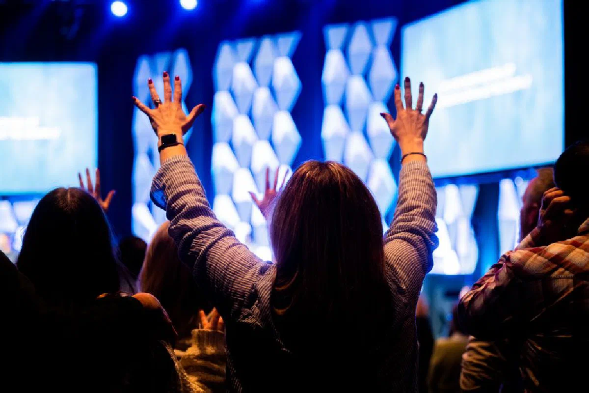 Energetic audience at an indoor concert with raised hands under bright stage lights.