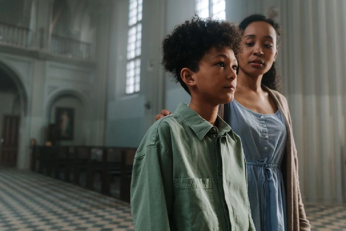 A mother and son stand in a serene church, reflecting contemplatively amidst the tranquil setting.