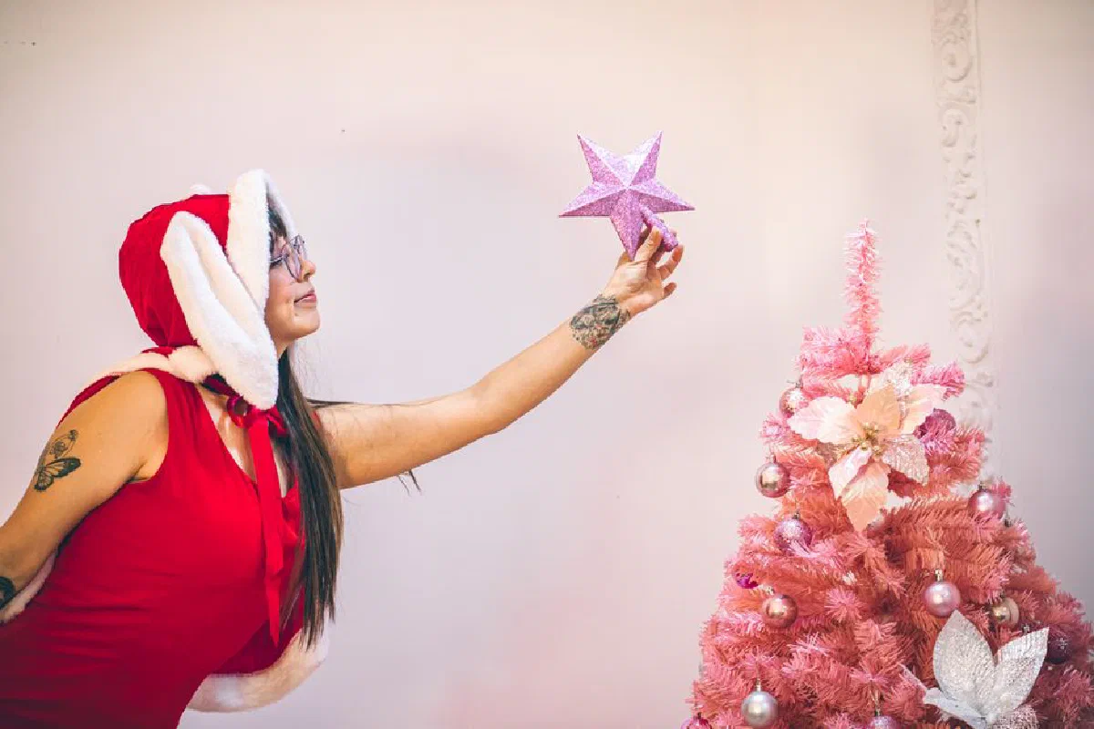 Woman in Santa outfit decorates pink Christmas tree with star indoors in Mexico City.