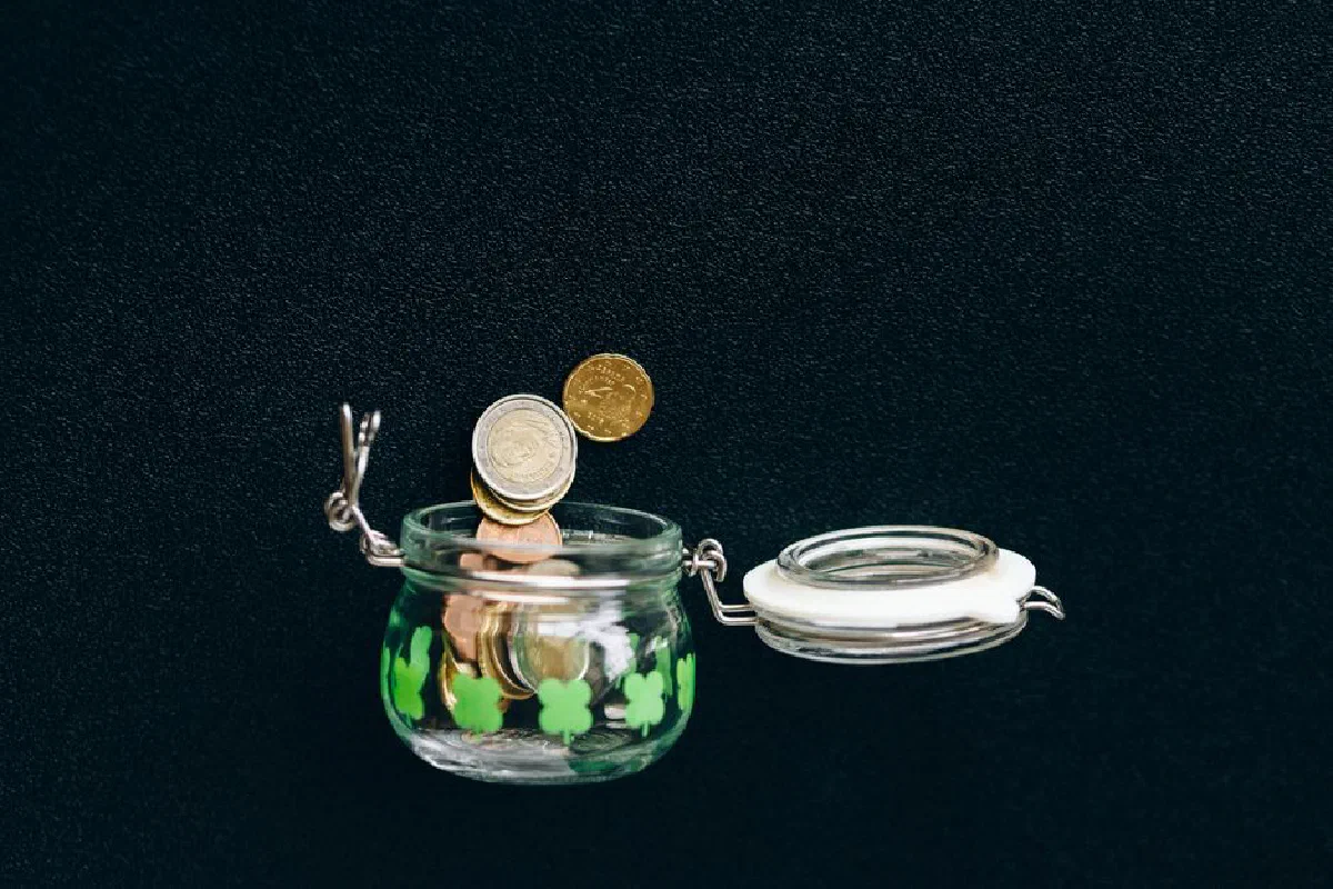 Glass jar with coins falling into it on a black background, symbolizing savings.