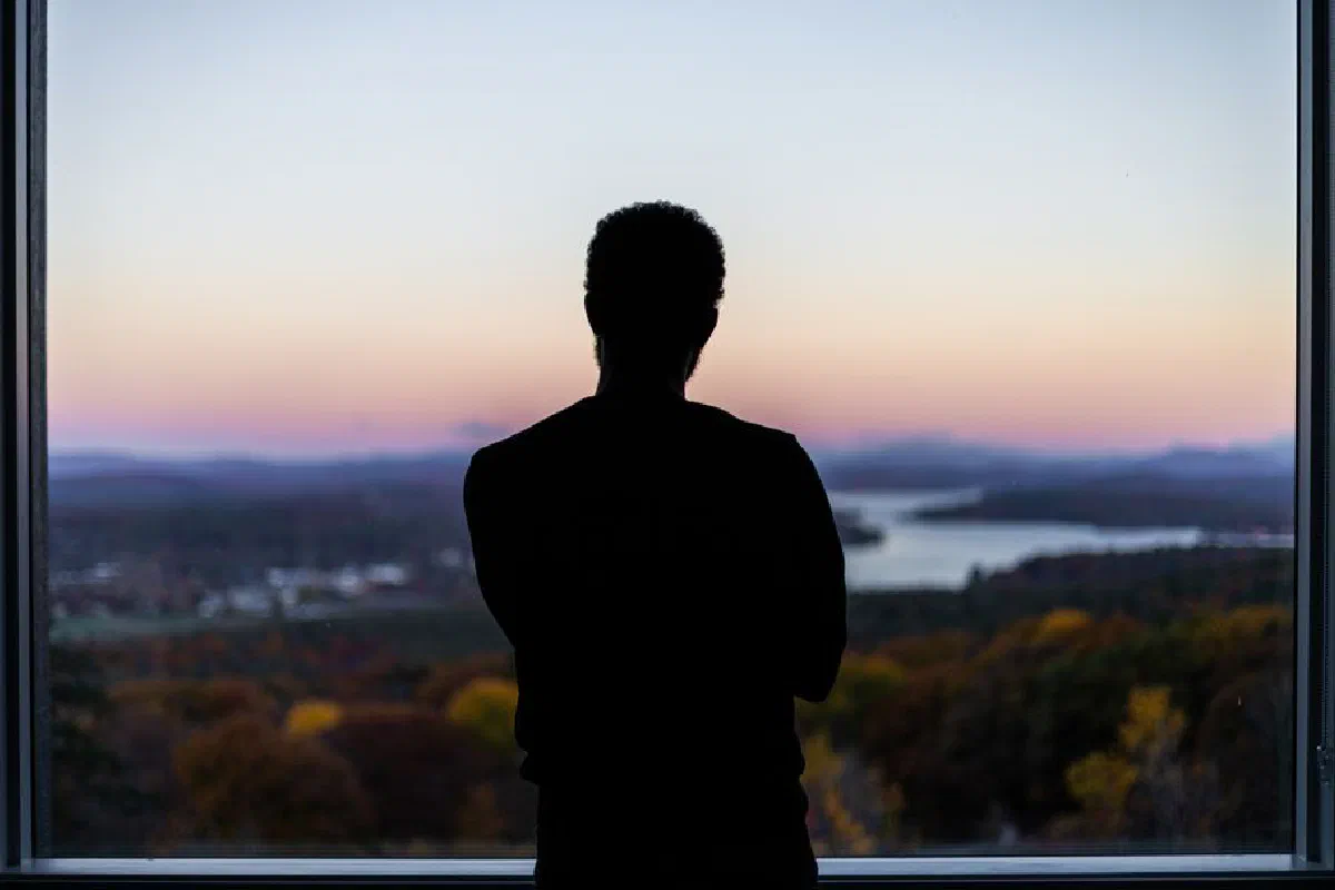 A silhouetted figure stands by a window, viewing a beautiful sunset landscape.