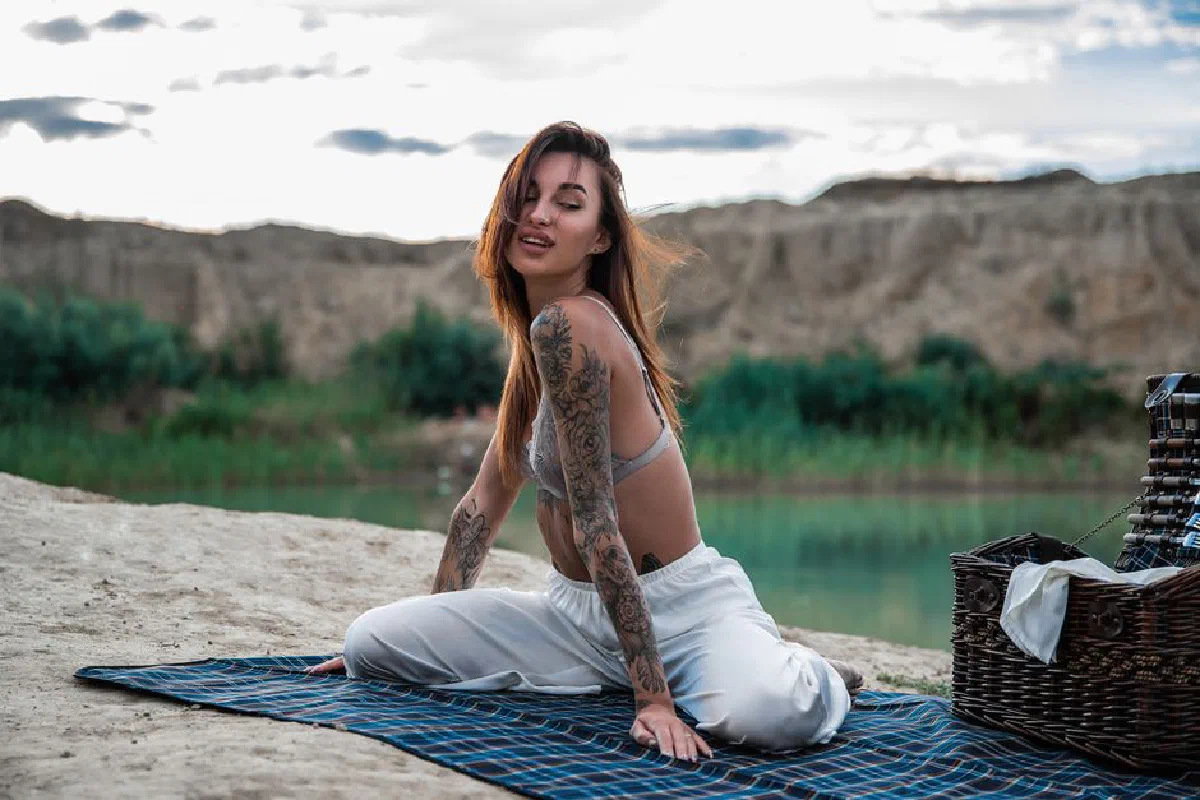 A young woman with tattoos enjoying a serene picnic by a lake on a sunny day.