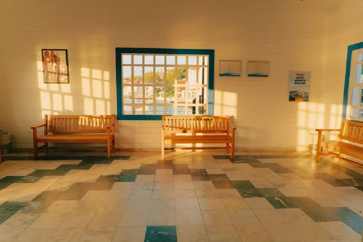 Warm sunlight filters through windows into a waiting room with wooden benches and tiled floor.