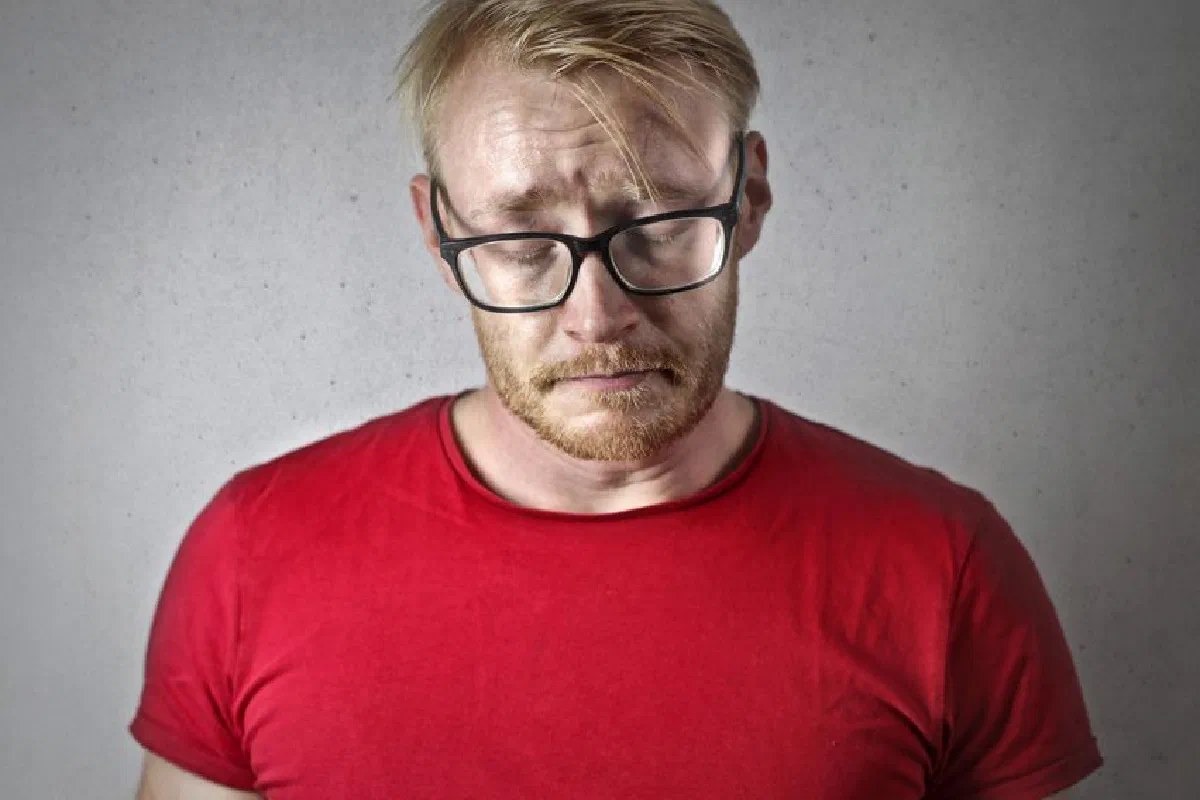 A bearded man wearing glasses looks down, expressing sadness against a gray background.