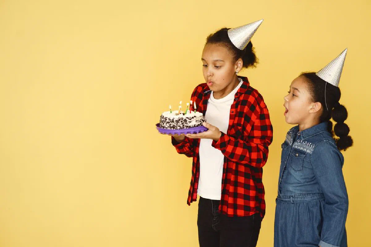 Two children in party hats enjoying a birthday celebration with a cake against a yellow backdrop.