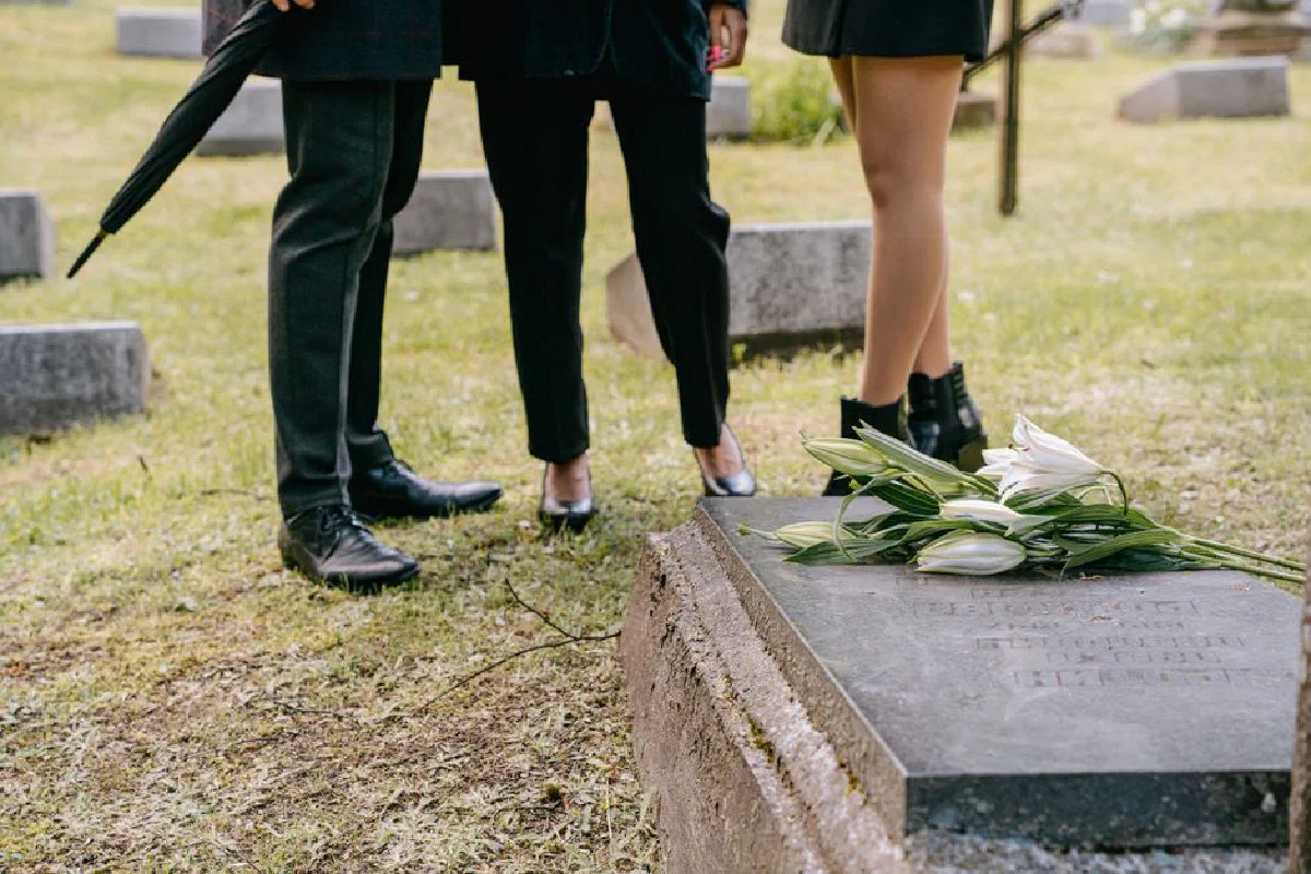 Two individuals in black standing by a grave with white lilies, evoking themes of remembrance.
