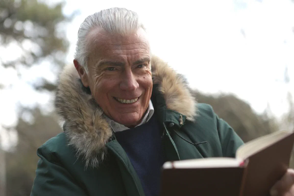 Senior man wearing a fur-lined coat, smiling while reading a book outdoors during winter.