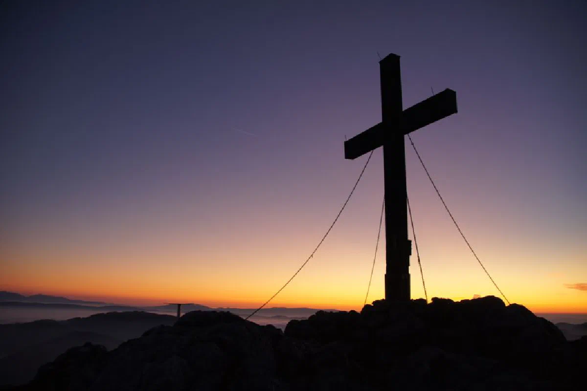 A dramatic cross stands silhouetted against a colorful sunset on a mountain peak, symbolizing faith and peace.