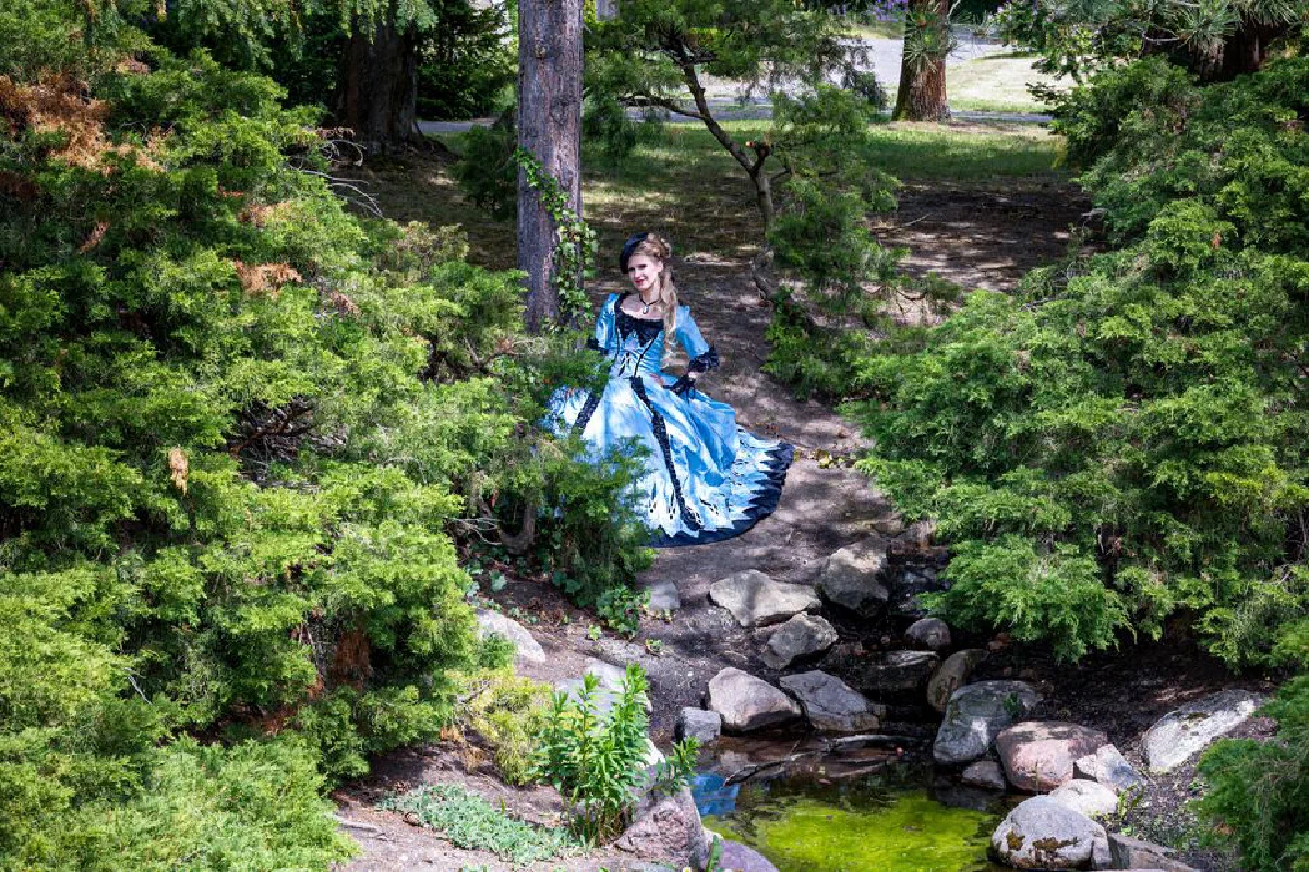 A woman dressed in a blue costume poses elegantly in a lush garden.
