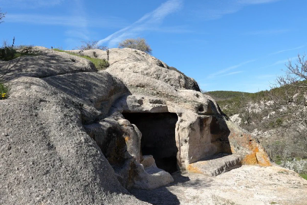 Explore an ancient rock cave nestled in a rugged natural landscape under a clear blue sky.
