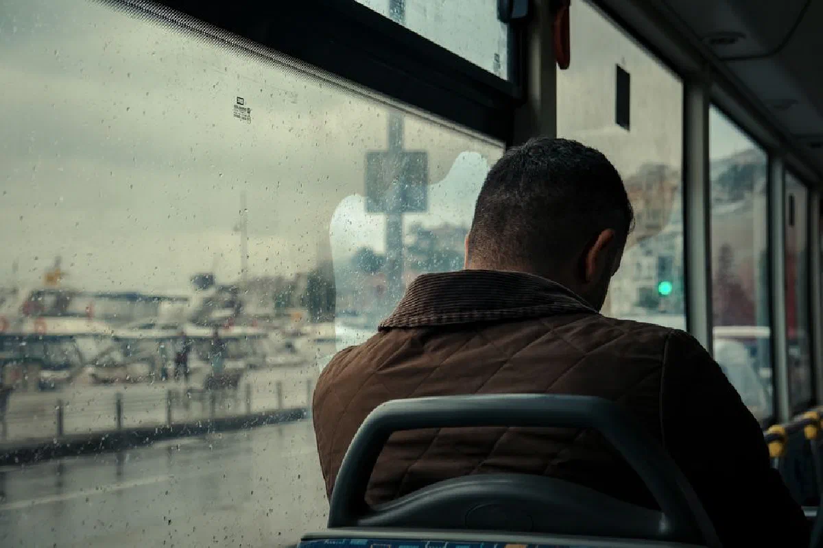 A solitary man on a bus gazes out at a rainy cityscape through a window.