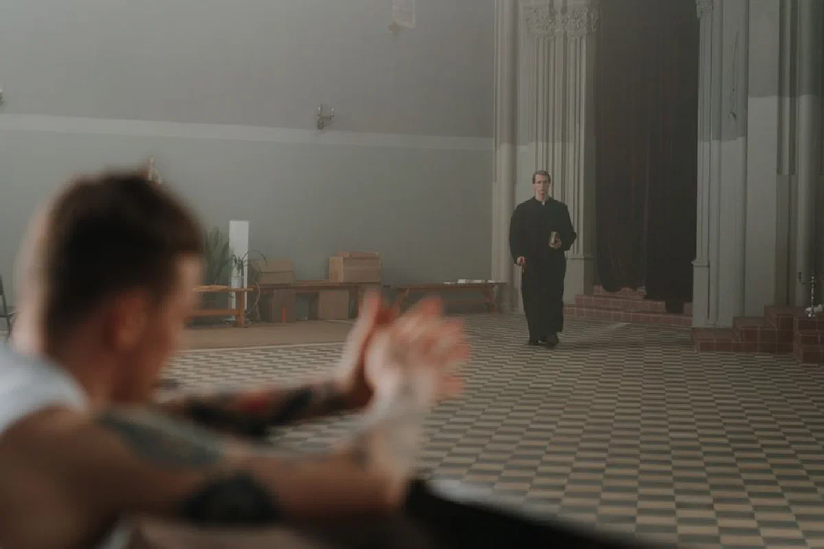 A tattooed man in a church with a priest approaching him, illustrating contrasting beliefs.