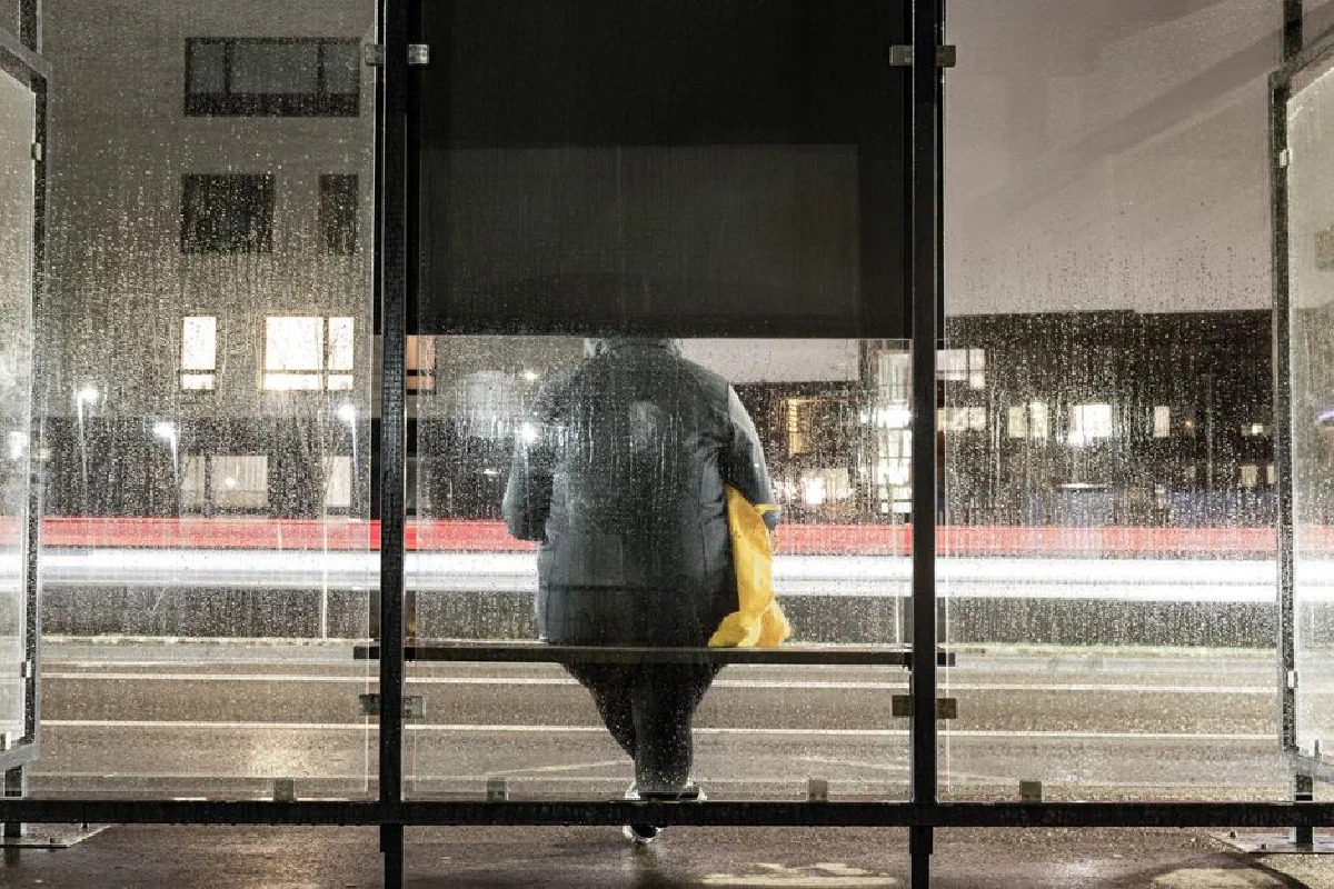 Person waiting at a rainy bus stop at night with light trails in the background.