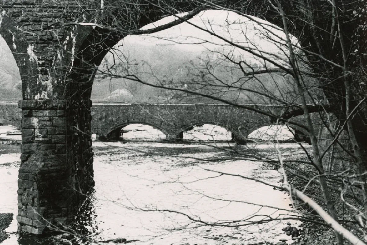 A sepia-toned image of a historic stone bridge over a partially frozen river in winter.
