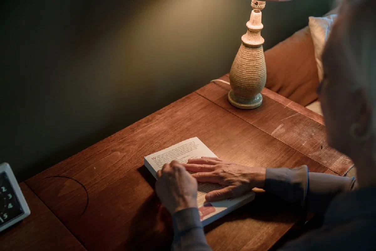 A senior woman reads a book on a wooden desk under warm lamp lighting.
