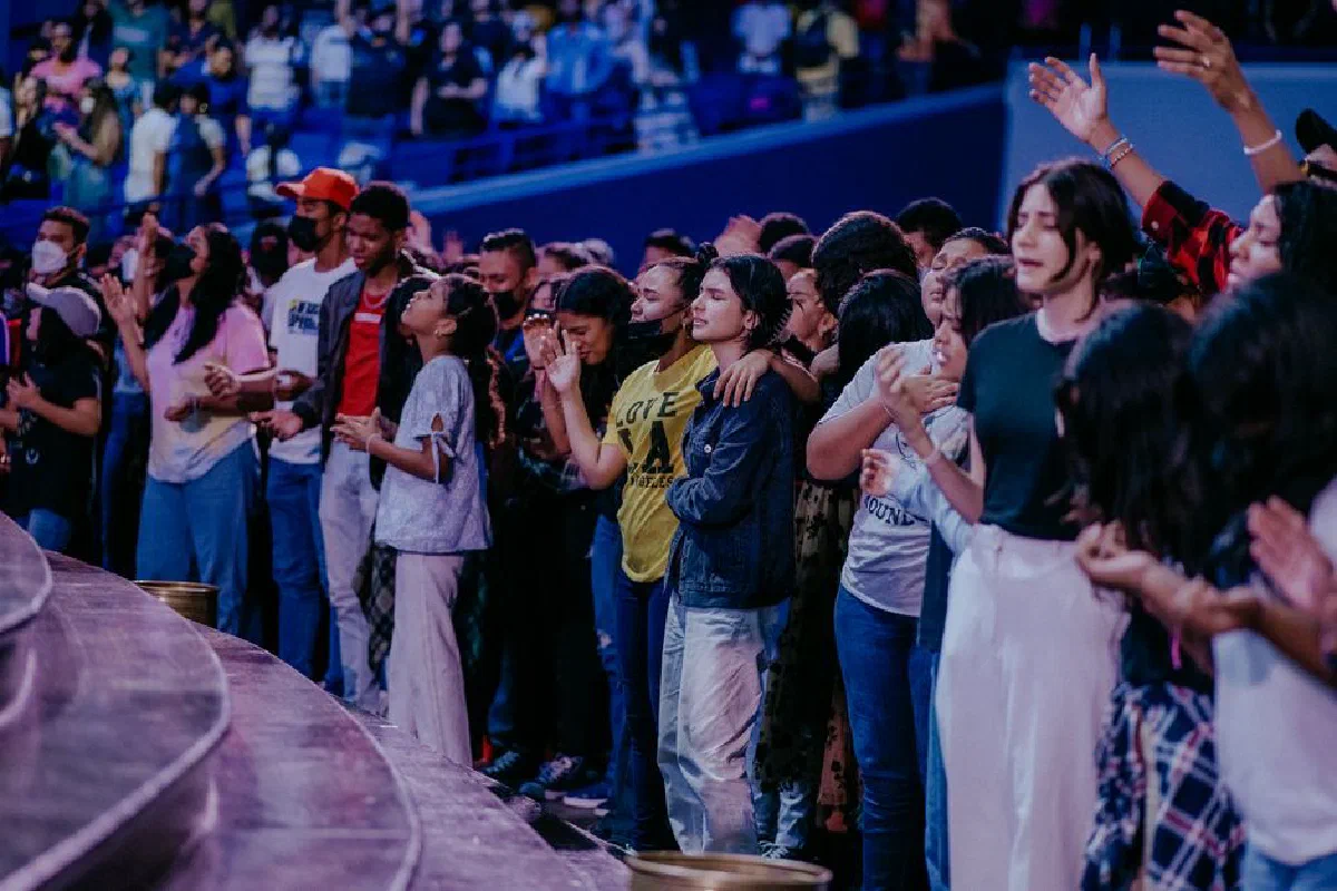 A congregation gathered indoors, raising hands and praying together in a spiritual setting.