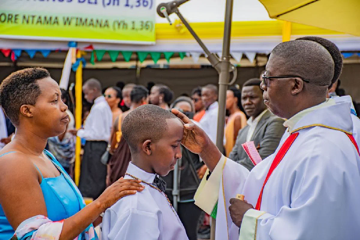 A boy receives a blessing during an outdoor religious ceremony with a priest, surrounded by family and guests.