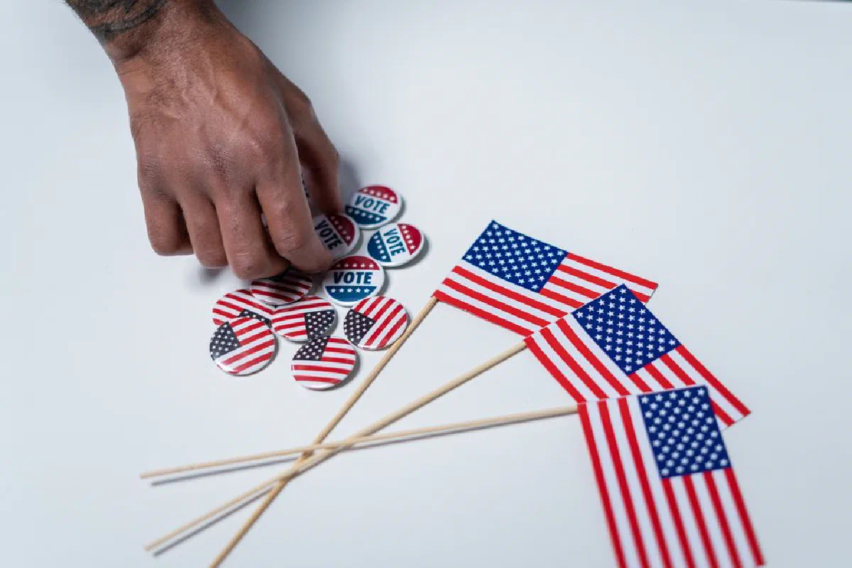 A hand reaches for voting buttons and American flags on a white background.