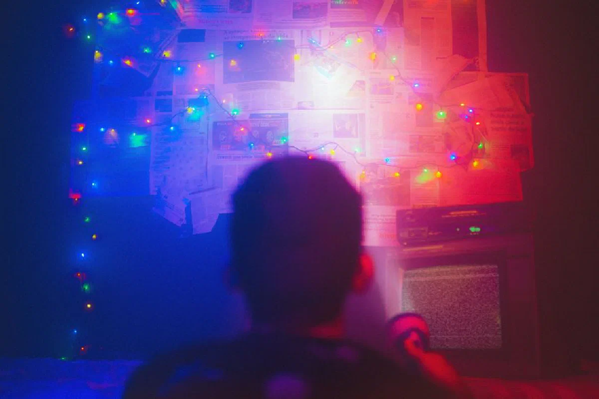 A dark room illuminated by colorful string lights with a wall of newspapers and TV static, creating a mysterious ambiance.