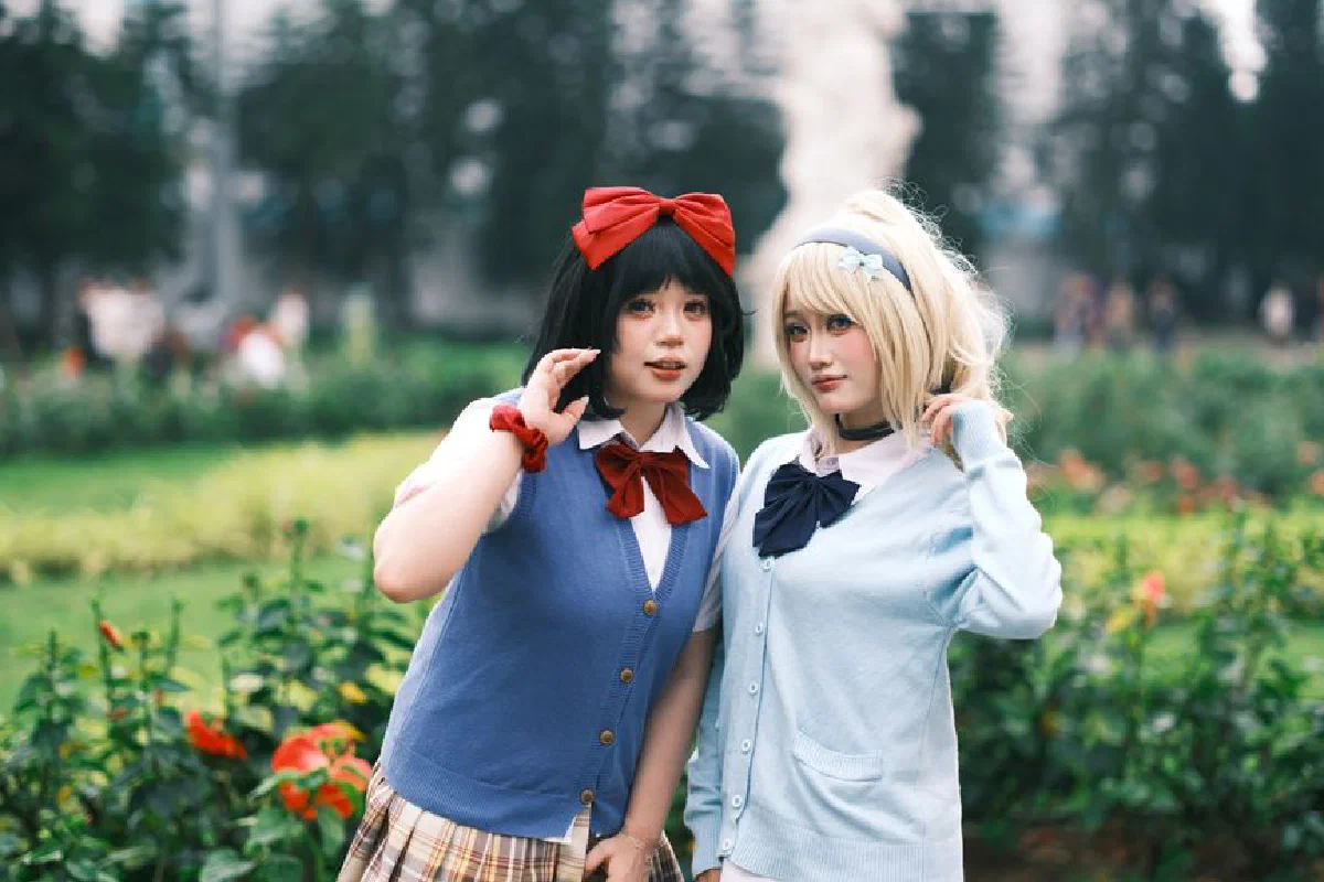 Two young women cosplay in school uniforms with vibrant bows, posing in a garden.