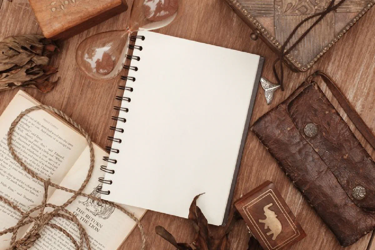A vintage-style flatlay with an open notebook, book, hourglass, and leather items on a wooden surface.
