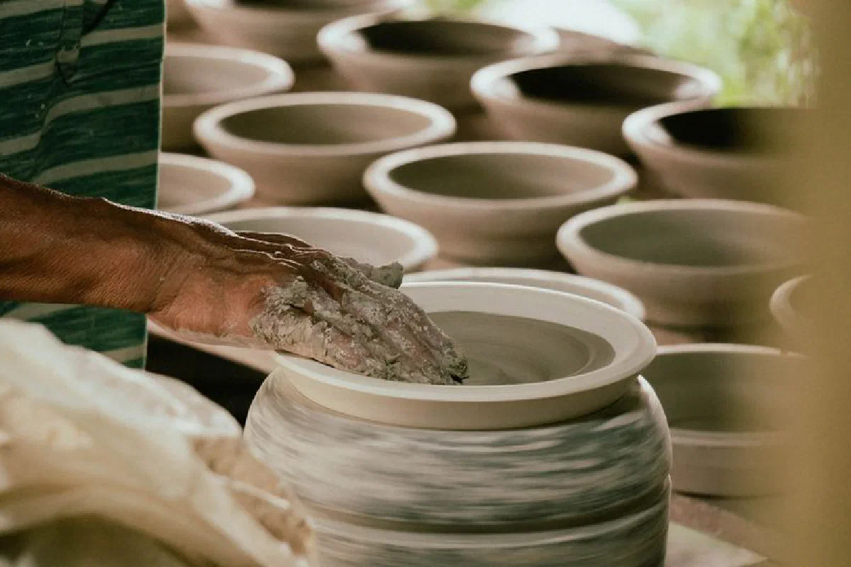 Hand shaping clay pot on a pottery wheel surrounded by finished pieces in a workshop.