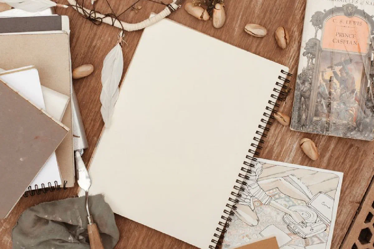 Flat lay of a sketchbook surrounded by artistic tools and a novel for creative inspiration.
