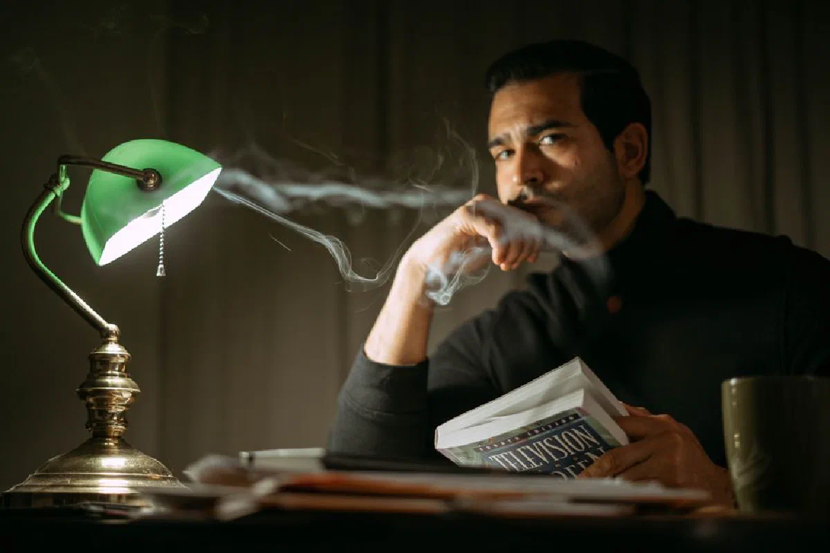 Focused young brunet wearing black sweater sitting at desk with books in dark home office while looking at camera and touching chin in contemplation