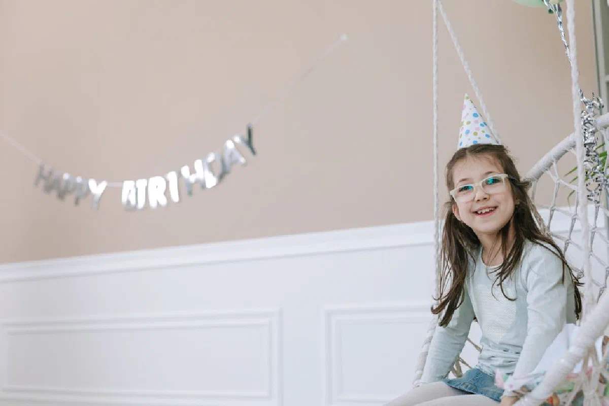 Young girl wearing glasses and party hat smiling on a swing indoors.