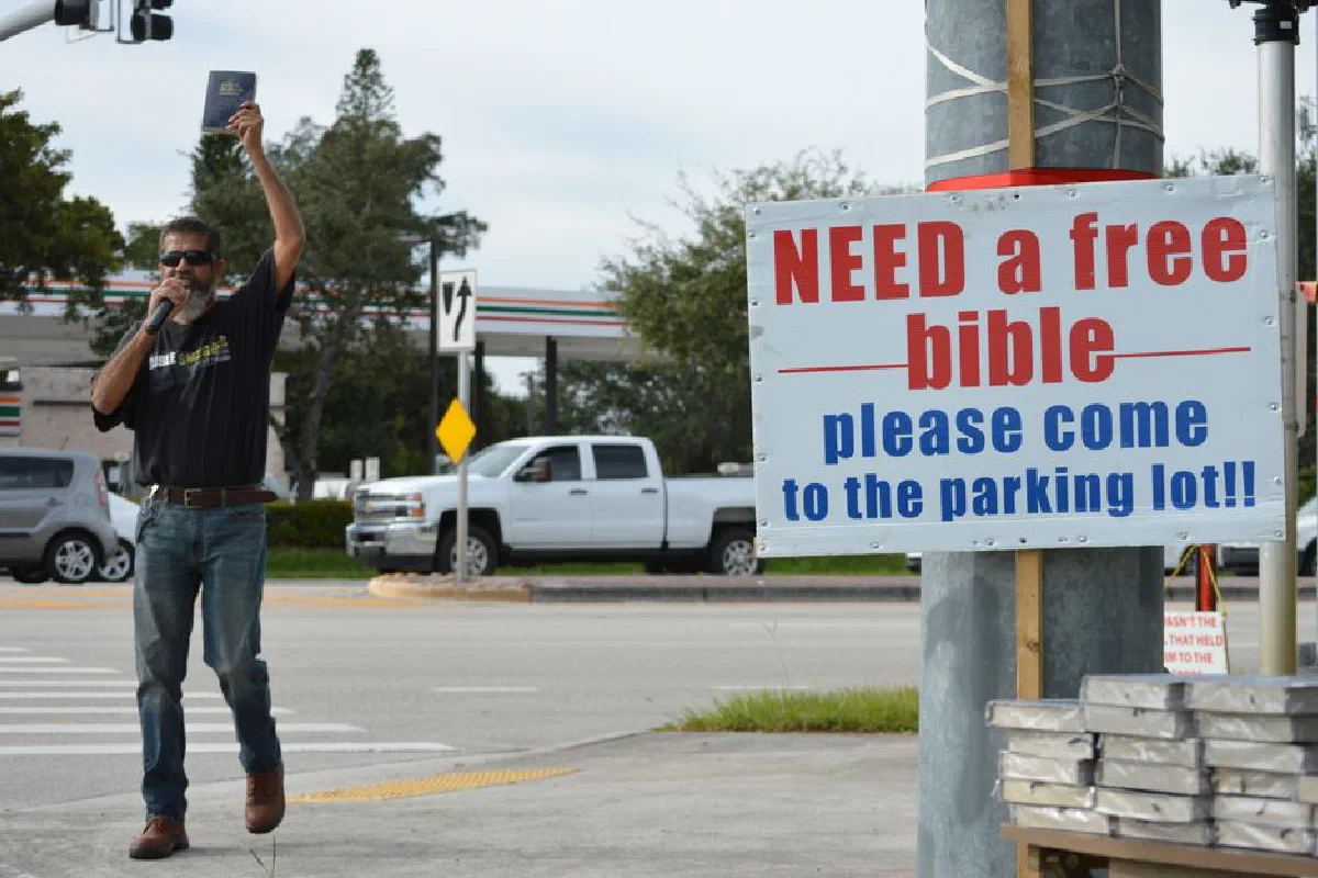 A man holding a Bible preaches outdoors with a sign offering free Bibles.