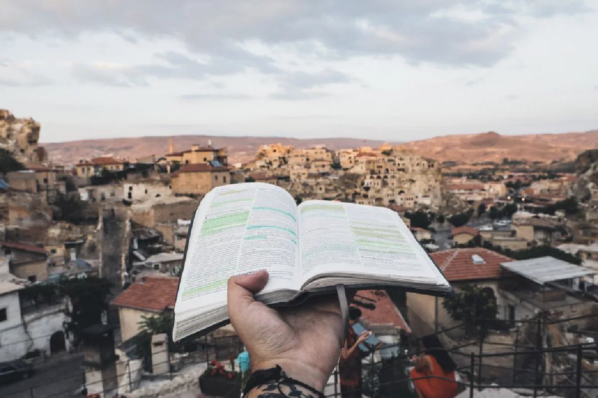 Hand holding an open Bible with a scenic view of ancient Cappadocia buildings in the background.