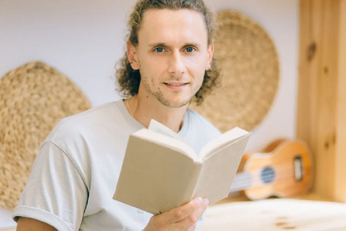 Adult male with curly hair reading a book indoors, smiling with a guitar in the background.