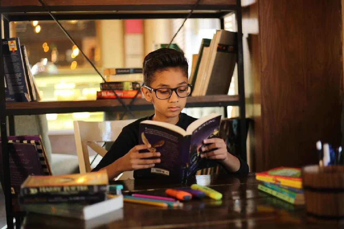 A young boy with glasses reading a book in a cozy library setting, surrounded by books and stationery.