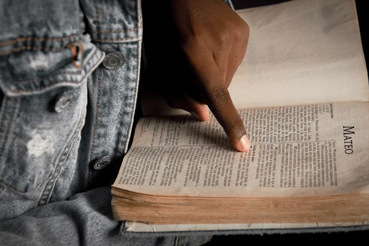 A person points to a passage in an old, open Bible, wearing a denim jacket.