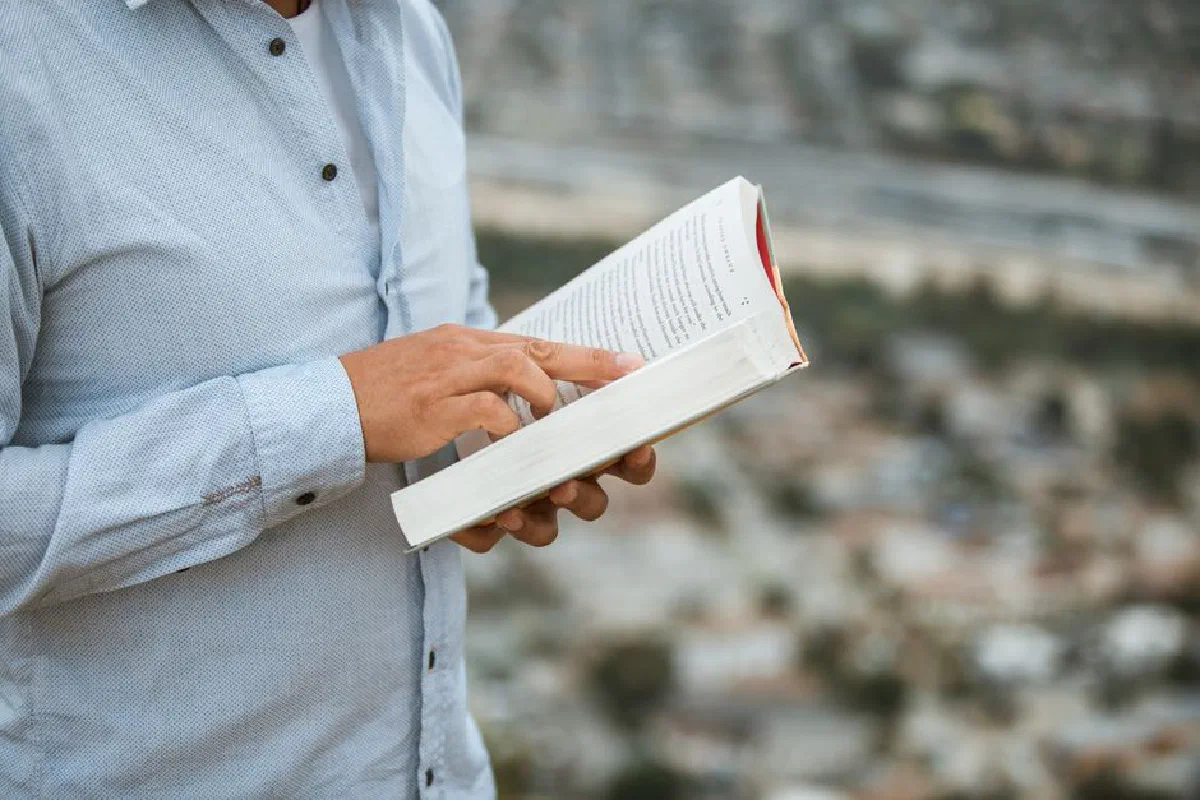 A person in a blue shirt reading a book, standing outdoors with a blurred cityscape in the background.