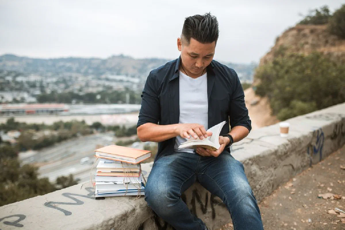 An adult man reads a book outdoors on a concrete wall with a stack of books beside him.