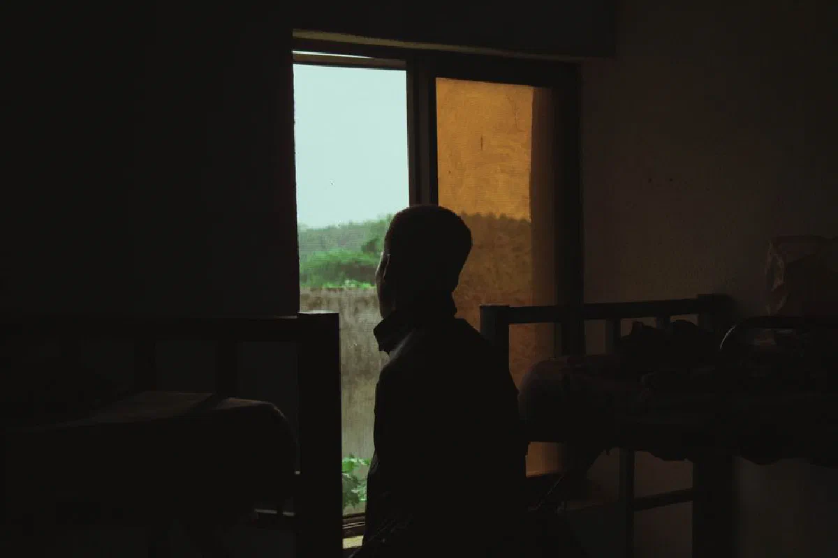 A man in silhouette stands by a window in a dimly lit room with bunk beds, evoking solitude.