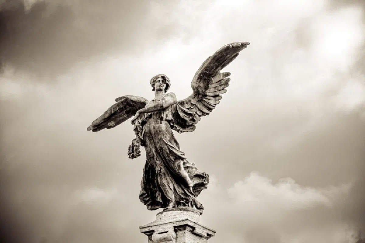 Sepia-toned image of an ancient angel statue with wings against a cloudy sky.