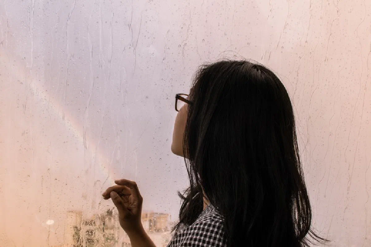 A woman gazes out a rain-speckled window at a faint rainbow, capturing a serene moment of reflection.