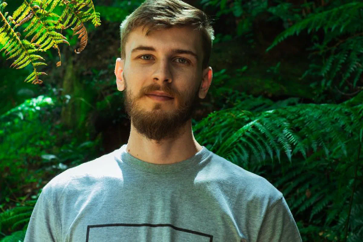 Portrait of a bearded man in a gray shirt, surrounded by lush green ferns in an outdoor setting.