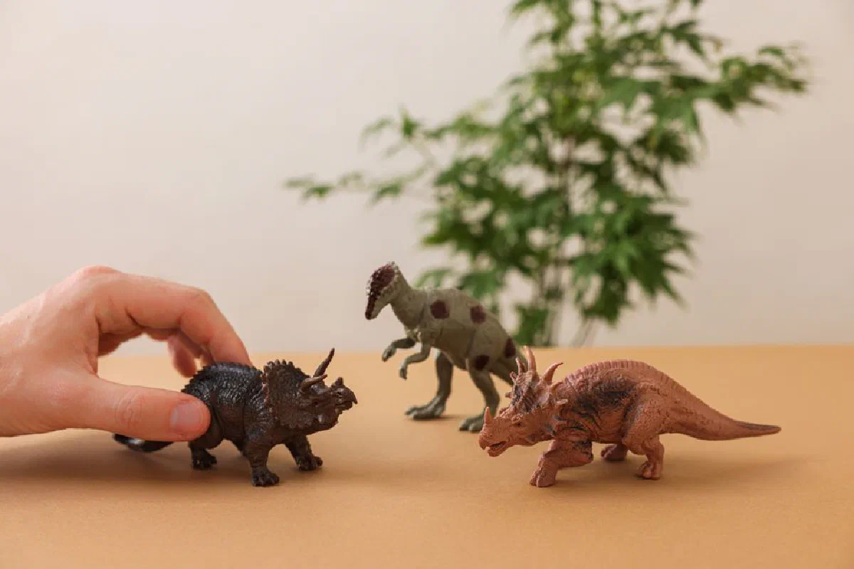 Hand arranging dinosaur figurines on a brown surface with greenery background.