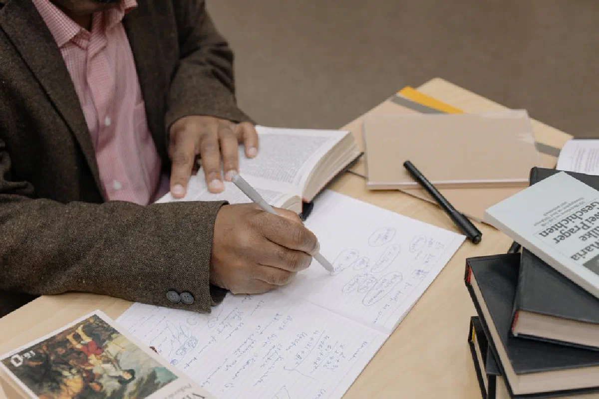 Man writing notes while researching with books at a desk indoors.