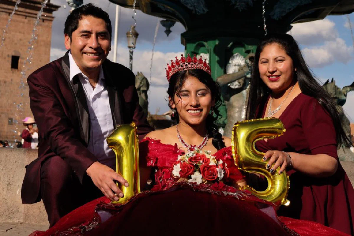A smiling Latina teenager celebrates her quinceañera with family under a sunny sky.