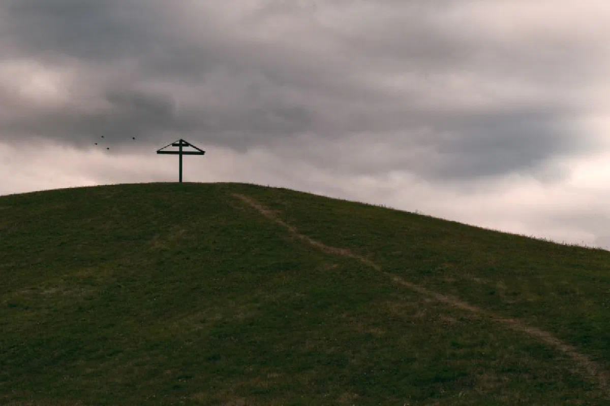 A solitary cross stands atop a grassy hill beneath a dramatic cloudy sky.