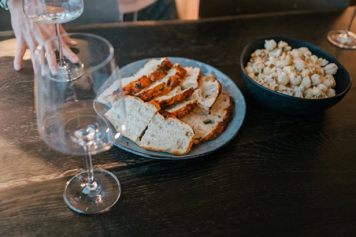 Sliced bread and popcorn in bowls on a dark wooden table, with wine glasses nearby.