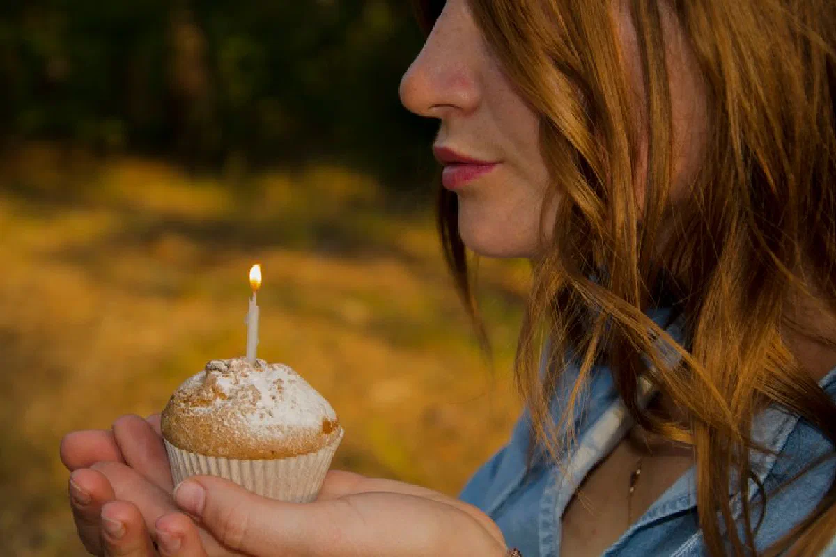 Close-up of a young woman holding a cupcake with a lit candle outdoors, suggesting a celebration.