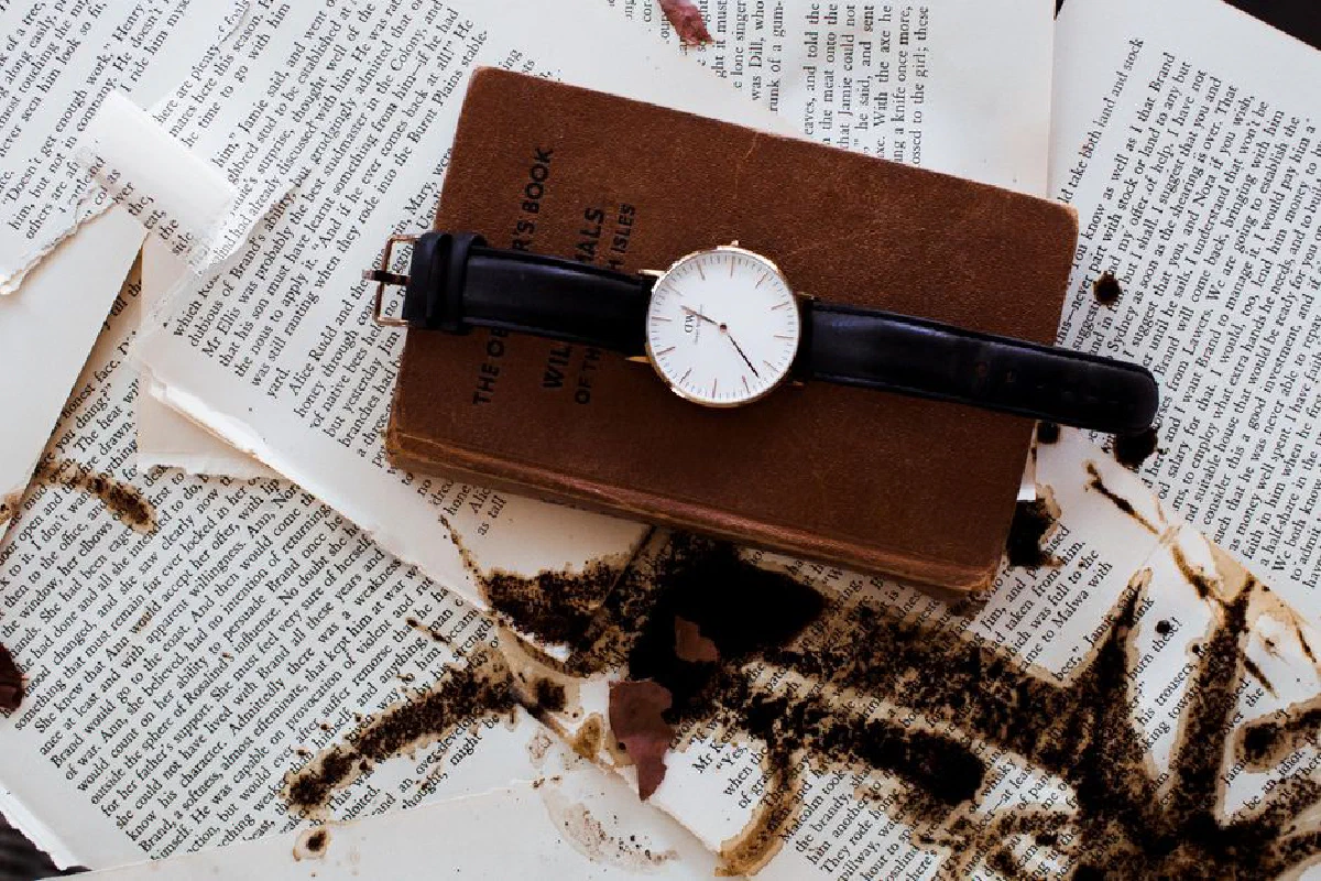 An analog wristwatch resting on a vintage book amid coffee-stained pages, evoking a timeless, literary feel.
