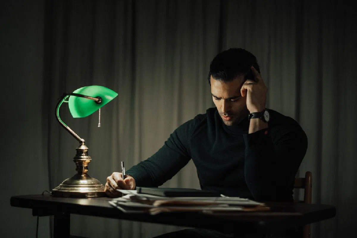 Concentrated male with pen taking notes while sitting at desk with documents and vintage lamp during work in dark room at home